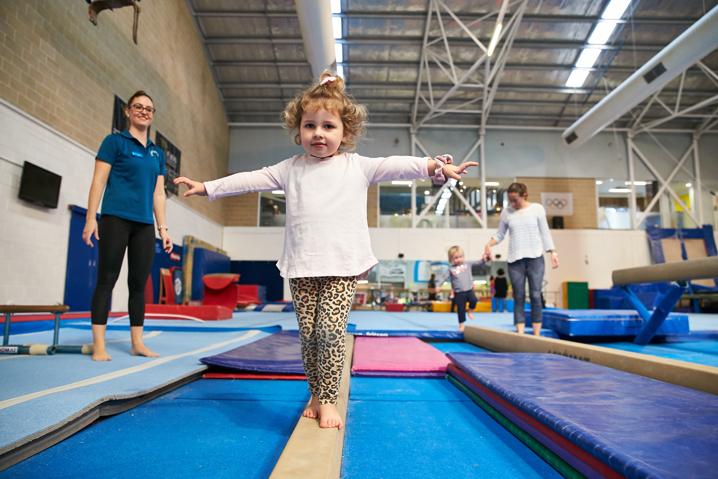 A child walking along a balance beam with a teacher looking on at the HBF Stadium Kids Gymnastics in Mount Claremont, Perth