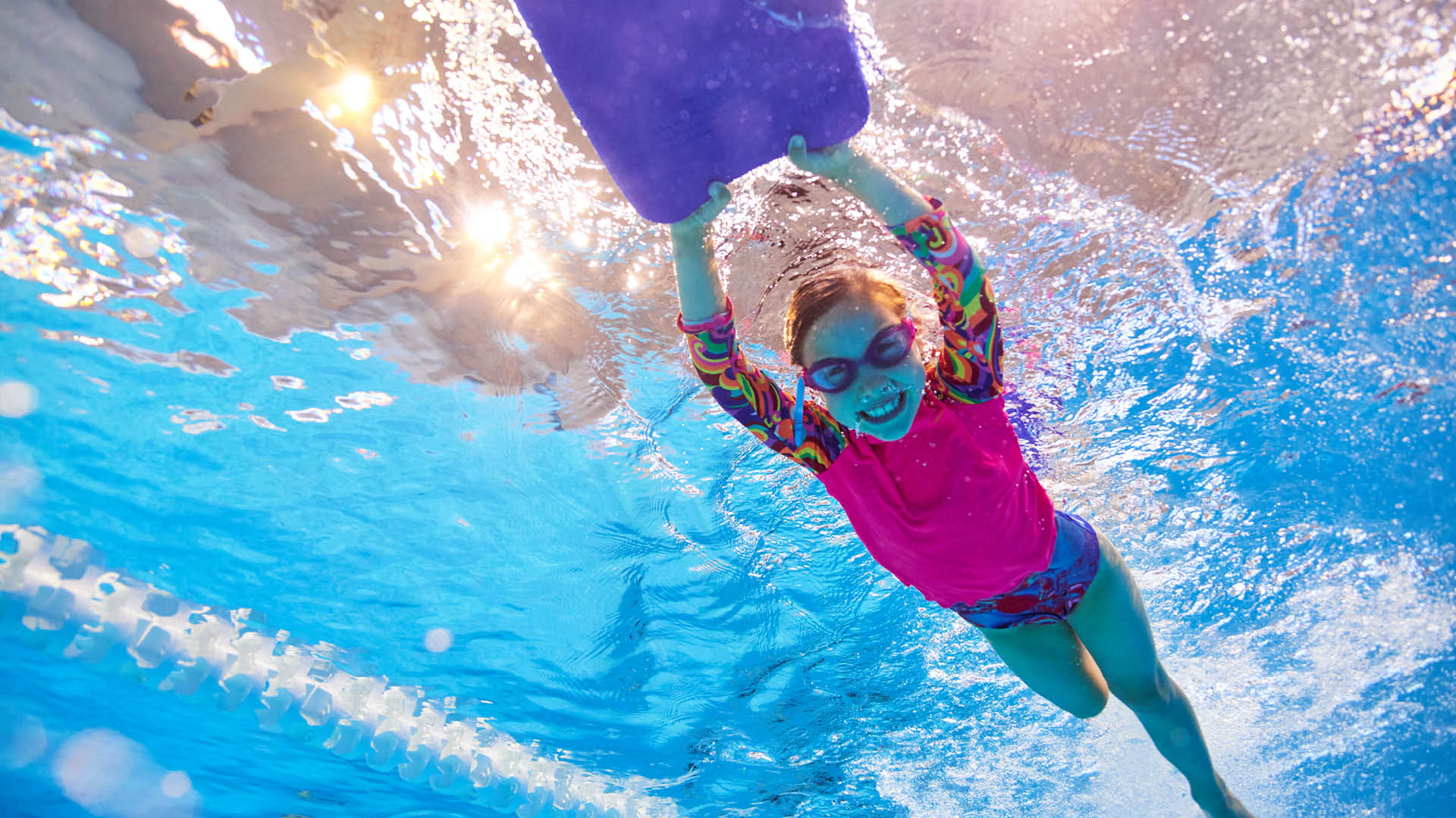 An underwater photo of a girl smiling as she swims along whilst holding at kick board at HBF Stadium in Mount Claremont
