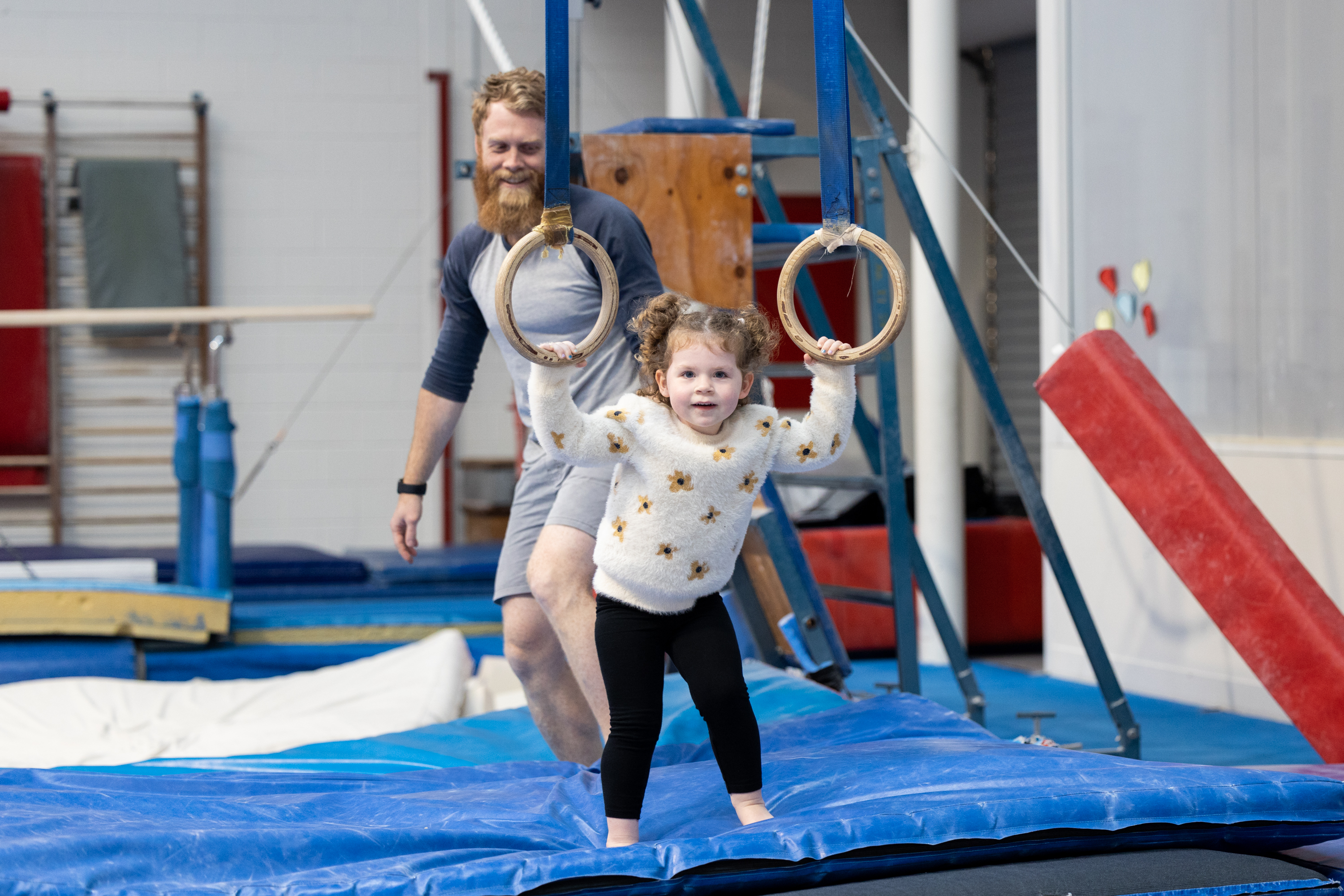 A child holding onto a pair of gymnastics stationary rings as her dad looks on at the HBF Stadium Kids Gymnastics in Mount Claremont, Perth
