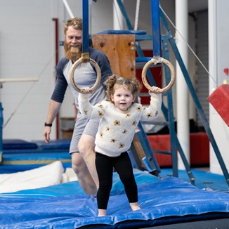 A child holding onto a pair of gymnastics stationary rings as her dad looks on at the HBF Stadium Kids Gymnastics in Mount Claremont, Perth