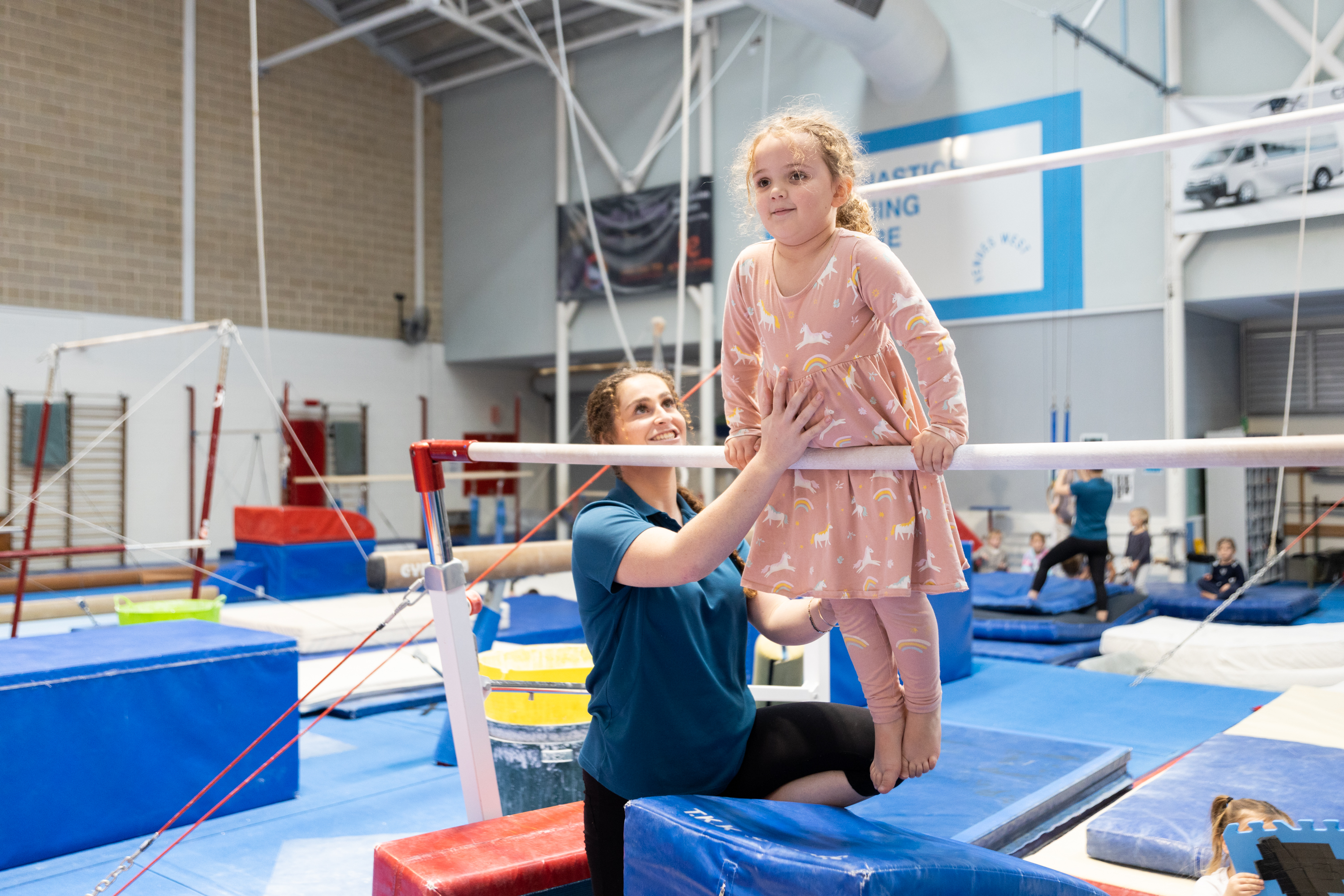 A child pulling herself up using a gymnastics bar with the assistance of a teacher at the HBF Stadium Kids Gymnastics in Mount Claremont, Perth