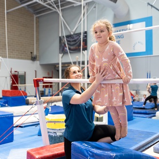 A child pulling herself up using a gymnastics bar with the assistance of a teacher at the HBF Stadium Kids Gymnastics in Mount Claremont, Perth