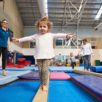 A child walking along a balance beam with a teacher looking on at the HBF Stadium Kids Gymnastics in Mount Claremont, Perth